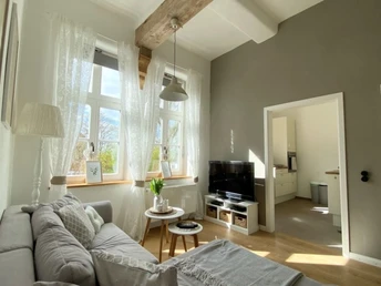 FeWo_Stiftsbrunnen_Wohnzimmer.png Helles Wohnzimmer mit Holzdecke, Sofa, Fernseher, Beistelltisch und Blick in die angrenzende Küche.