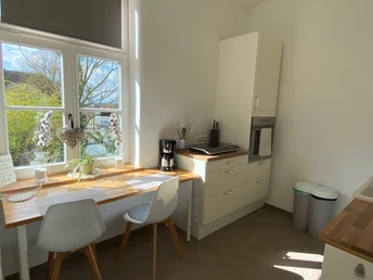 FeWo_Stiftsbrunnen_Küche.png Helle Küche mit Fenster, kleinem Tisch und zwei Stühlen, ausgestattet mit moderner Kaffeemaschine und Backofen.