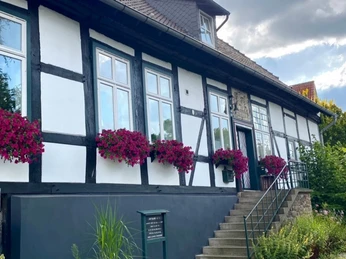 Half-timbered house with white walls, red flowers on the windows, stairs lead to the entrance.