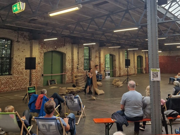 Herford- Mein Sommer Meine Stadt-Pro Herford GmbH 3.jpeg In einer alten Fabrikhalle sitzen Menschen auf Liegestühlen und lauschen einem Vortrag oder einer Lesung.