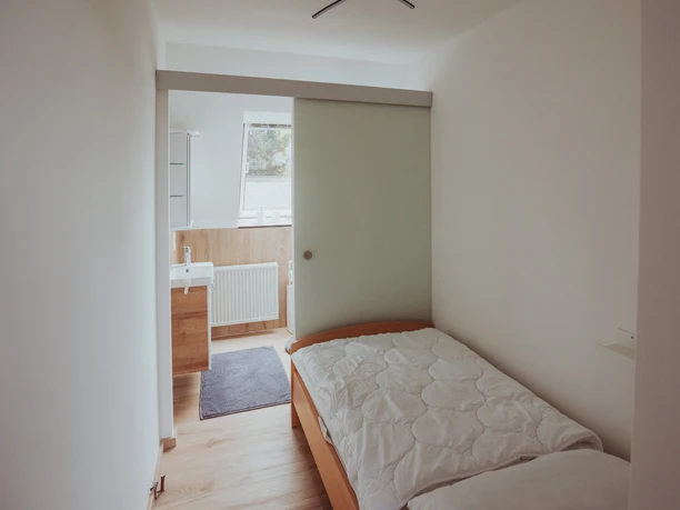 FeWo Hille Krysiak Ein Einzelzimmer mit Holzbett und Tageslicht, angrenzendem Badezimmer mit Waschbecken und Spiegel.