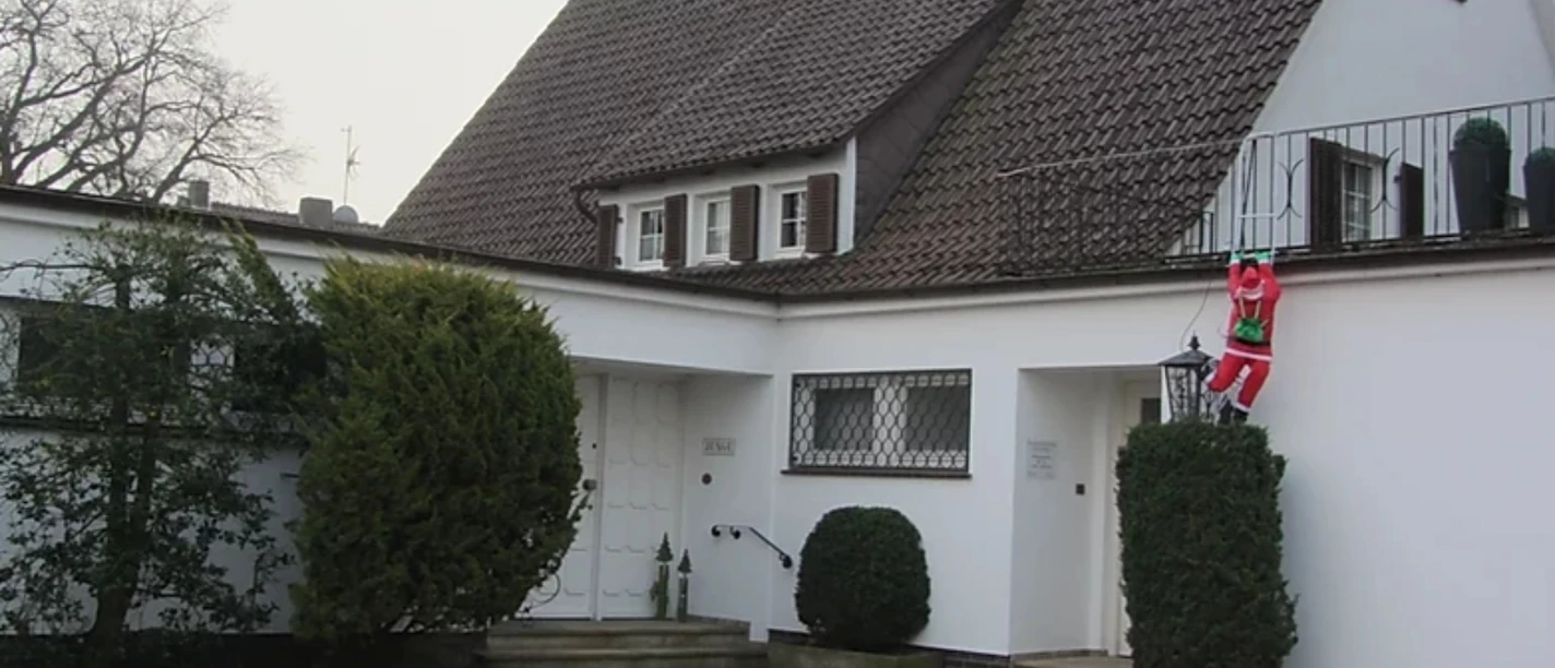 Apartment_Stemwede.jpg Gemütliches weißes Haus mit Dachschindeln und einem Weihnachtsmann, der an einer Hecke klettert.