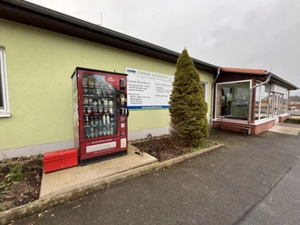Proviantomat Standort Struppen Landfleischerei Roter Verkaufsautomat mit Lebensmitteln vor einem hellgrünen Gebäude, daneben ein Schild mit Angeboten und eine kleine, konische Tanne.