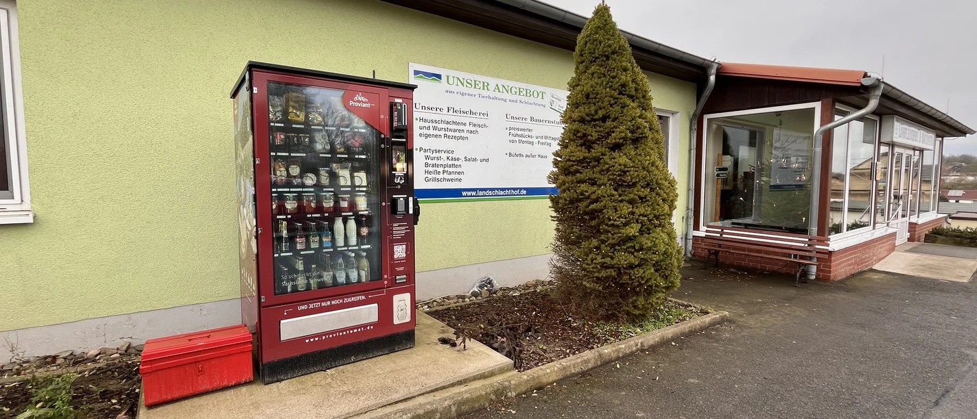 Proviantomat Standort Struppen Landfleischerei Roter Verkaufsautomat mit Lebensmitteln vor einem hellgrünen Gebäude, daneben ein Schild mit Angeboten und eine kleine, konische Tanne.