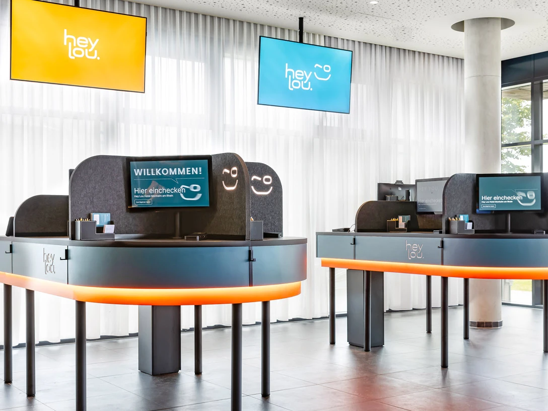 Rezeption im Hey Lou Monheim am Rhein Moderne Rezeption mit zwei Servicepunkten, beleuchtetem Tresen, digitalen Bildschirmen und hellen Vorhängen.
