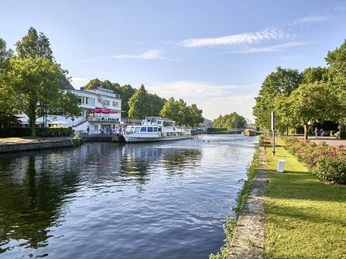 Wasserbahnhof (2017), Mülheim an der Ruhr Passagierschiff auf ruhigem Fluss neben grüner Anlage und einem Gebäude in Mülheim.