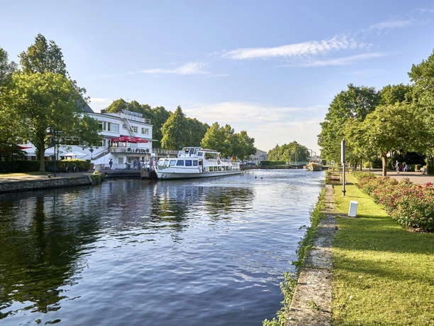 Wasserbahnhof (2017), Mülheim an der Ruhr Passagierschiff auf ruhigem Fluss neben grüner Anlage und einem Gebäude in Mülheim.