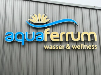 aquaferrum aquaferrum-Logo auf grauer Gebäudefassade, begleitet von der Aufschrift „wasser & wellness“.aquaferrum logo on gray building facade, accompanied by the inscription "water & wellness".aquaferrum-logo på grå bygningsfacade, ledsaget af påskriften "water & wellness".aquaferrum-logo op de grijze gevel van het gebouw, met het opschrift "water & wellness".
