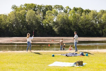 Naturbadesee Mindenerwald Familie spielt am See auf einer grünen Wiese; im Hintergrund ein ruhiger Natursee.