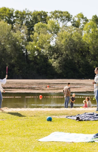 Naturbadesee Mindenerwald Familie spielt am See auf einer grĂĽnen Wiese; im Hintergrund ein ruhiger Natursee.