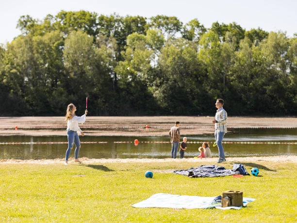 Naturbadesee Mindenerwald Familie spielt am See auf einer grünen Wiese; im Hintergrund ein ruhiger Natursee.