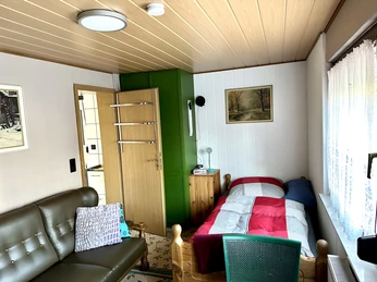 Helles Wohnzimmer mit Holzmöbeln, grünem Schrank, Einzelbett, Couch und natürlichem Lichteinfall.