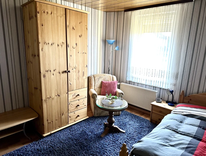 Ein gemütliches Schlafzimmer mit Holzschrank, Sessel, Teppich und Fenster, das Tageslicht hereinlässt.