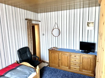 Schlafzimmer mit Einzelbett, Ledersessel, Kommode mit flachem Fernseher und gestreifter Tapete.