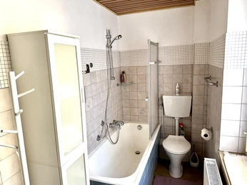 Bad mit Badewanne, Dusche, WC und Fliesen. Weiße Möbel und Handtuchhalter. Helle Wandgestaltung.