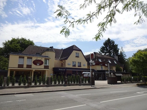 Außenansicht vom Hotel im Park, Haan Das Bild zeigt die Außenansicht eines Hotels mit braunem Dach und mehreren Stockwerken in einer städtischen Umgebung, umgeben von Bäumen und einer Straße im Vordergrund.