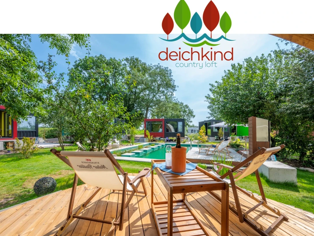 Deichkind Country Loft Zwei Liegestühle und ein Tisch mit Weinkühler auf einer Holzterrasse, umgeben von grüner Natur.