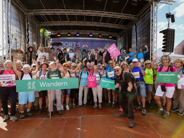 06_30 Sports United (c) sazinc.jpg Eine Gruppe von Menschen steht lächelnd vor einer Bühne mit bunten Schildern in der Hand.