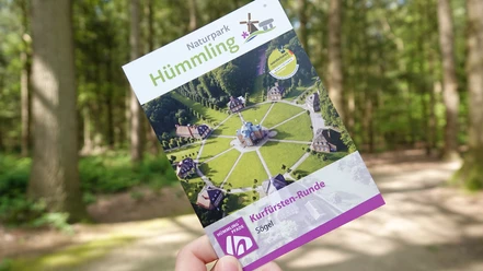 Hümmling-Pfade Kurfürsten-Runde – Schnappschuss Clemenswerth ©Emsland Tourismus GmbH (6).jpg Eine Hand hält einen Flyer der Kurfürsten-Runde im Naturpark Hümmling auf einem Waldweg.