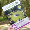 Hümmling-Pfade Kurfürsten-Runde – Schnappschuss Clemenswerth ©Emsland Tourismus GmbH (6).jpg Eine Hand hält einen Flyer der Kurfürsten-Runde im Naturpark Hümmling auf einem Waldweg.