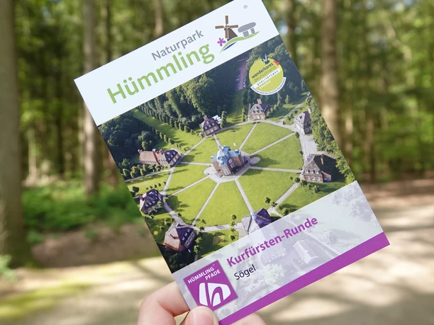 Hümmling-Pfade Kurfürsten-Runde – Schnappschuss Clemenswerth ©Emsland Tourismus GmbH (6).jpg Eine Hand hält einen Flyer der Kurfürsten-Runde im Naturpark Hümmling auf einem Waldweg.