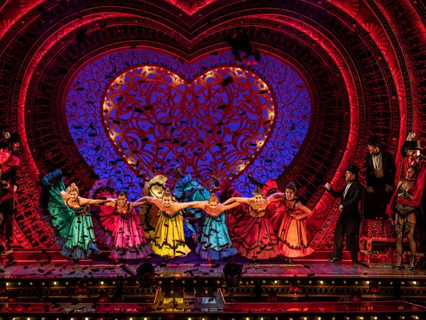 Moulin Rouge! The musical On an elaborately lit stage set, performers dance lively can-can style in front of a large, heart-shaped backdrop. The costumes are colorful and detailed, and the performance conveys energy and movement.