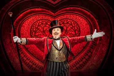 Moulin Rouge! Das Musical Ein strahlender Schauspieler im eleganten, roten Frack, mit Zylinder und Stock, vor leuchtendem Herzmotiv.