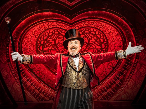 Moulin Rouge! The musical A radiant actor in an elegant red tailcoat, with top hat and cane, in front of a glowing heart motif.