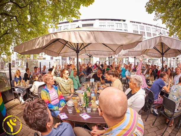 SC Janus Queer Games Brunch Large group at an open brunch under parasols, many raise their hands together.