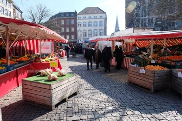 Besucher*innen auf einem lebhaften Wochenmarkt, eingerahmt von bunten Marktständen, vor historischer Kulisse.