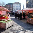 Besucher*innen auf einem lebhaften Wochenmarkt, eingerahmt von bunten Marktständen, vor historischer Kulisse.