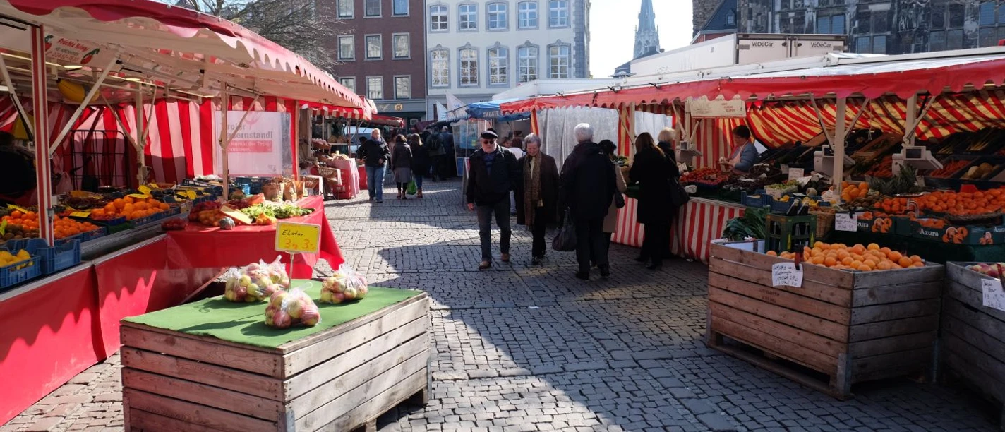 Besucher*innen auf einem lebhaften Wochenmarkt, eingerahmt von bunten Marktständen, vor historischer Kulisse.