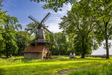 Hüvener Mühle – Radfahren Hümmling ©Emsland Tourismus GmbH (2).jpg Zwei Radfahrende auf einem grünen Weg neben der historischen Hüvener Mühle im Emsland.