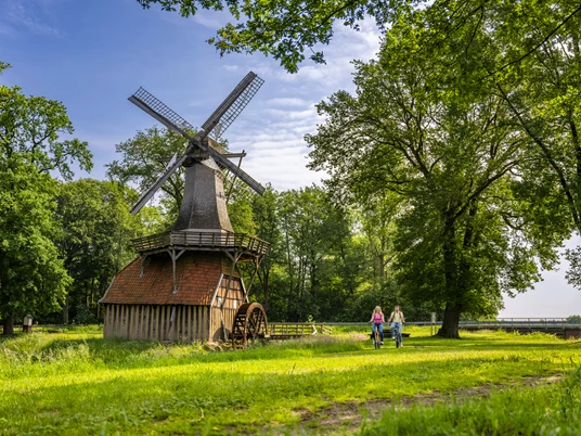 Hüvener Mühle – Radfahren Hümmling ©Emsland Tourismus GmbH (2).jpg Zwei Radfahrende auf einem grünen Weg neben der historischen Hüvener Mühle im Emsland.