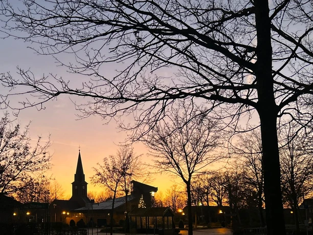Radrundtour auf dem Hümmling ©Birgit Kuper-Gerdes (17).jpg Kirche mit erleuchtetem Turm im Abendlicht, umgeben von Bäumen vor farbigem Himmel bei Sonnenuntergang.