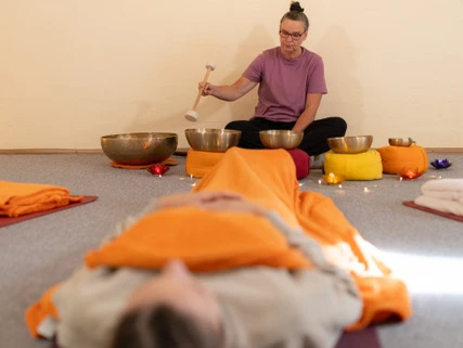 Klang- / Entspannungsreise Frau spielt Klangschalen neben liegender Person, sanftes Licht und warme Farben schaffen Ruhe.