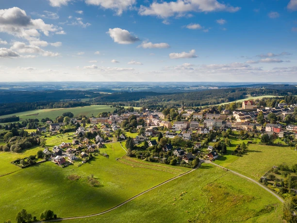 Aerial view of Schöneck in summer, surrounded by green meadows and forests under a blue sky.