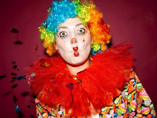 Karneval Clown Ein Clown mit bunter Perücke, rotem Kragen und Luftschlangen auf dunkelrotem Hintergrund.