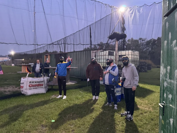 IMG_5258.jpeg Gruppe von Spielern mit Schutzmasken auf einem beleuchteten Paintballfeld bei Abenddämmerung