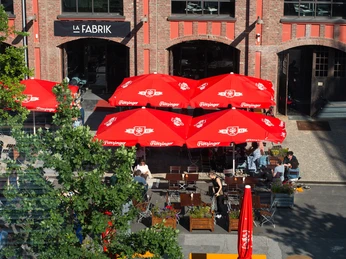 La Fabrik (2) Außengastronomie mit Schirmen und mehreren Sitzgruppen. Dahinter befindet sich ein Gebäude aus Ziegelsteinen, auf dem "La Fabrik" steht.