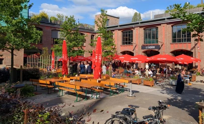 La Fabrik (1) Außenterrasse mit roten quadratischen Schirmen und Sitzgruppen, die von einigen Menschen besucht sind. Dahinter befindet sich ein großes Gebäude aus Ziegelsteinen.