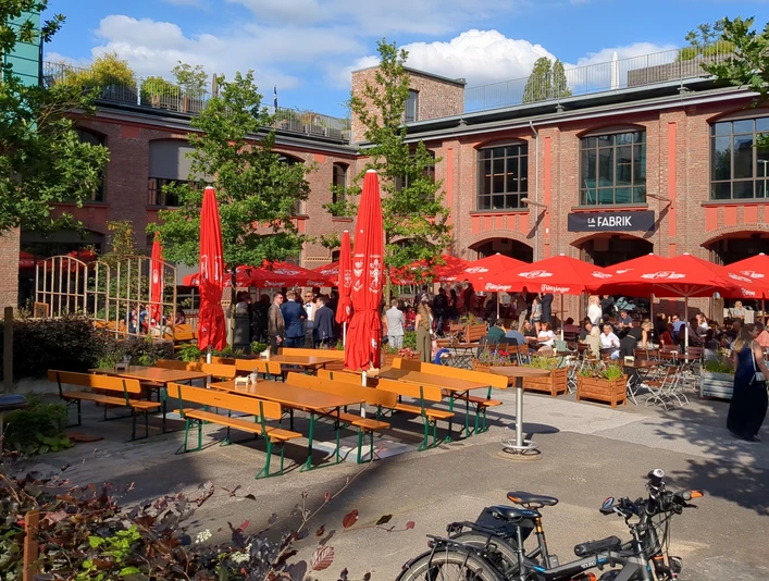 La Fabrik (1) Außenterrasse mit roten quadratischen Schirmen und Sitzgruppen, die von einigen Menschen besucht sind. Dahinter befindet sich ein großes Gebäude aus Ziegelsteinen.