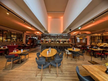 La Fabrik (4) Innenraum mit mehreren Sitzgruppen aus teils runden, teils quadratischen Tischen. In der Mitte befindet sich eine Bar