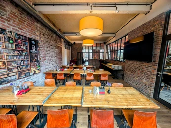 Innenraum mit Steinwänden, großen Deckenleuchten und drei lange Tische mit Stühlen, die sich hintereinander befinden.