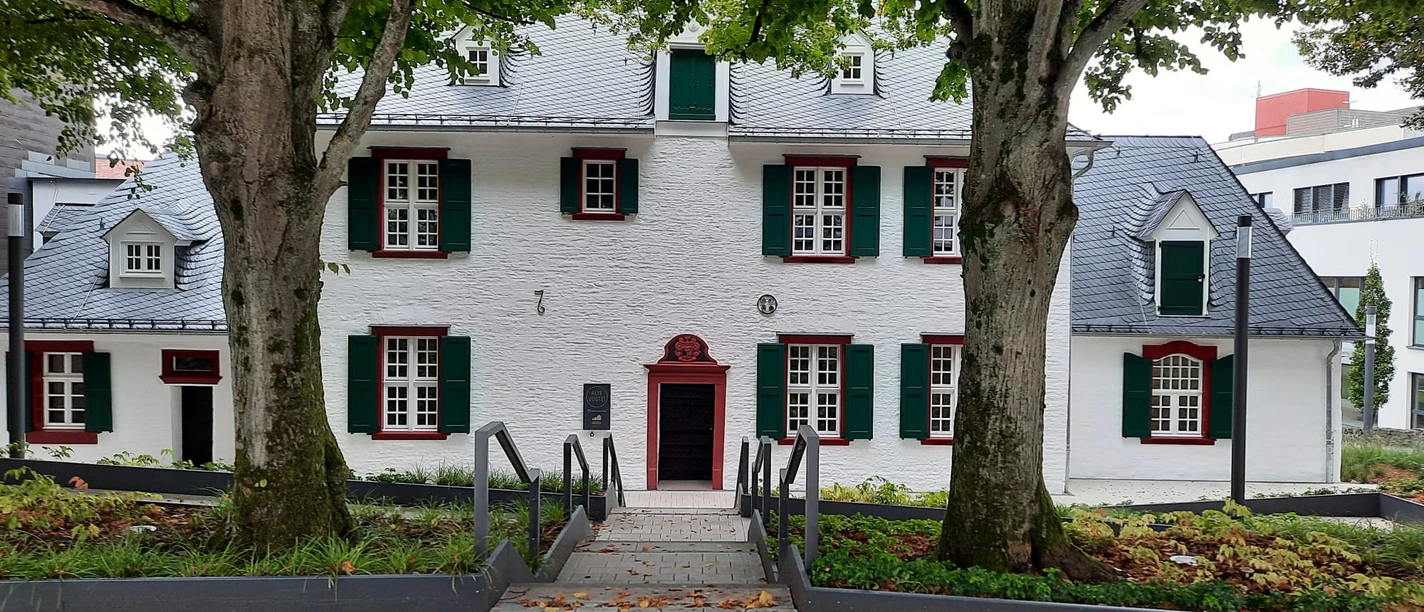 Foto Vogtei Gummersbach.jpg Historisches, weißes Gebäude mit Schindeldach, grünen Fensterläden, umgeben von zwei großen Bäumen.