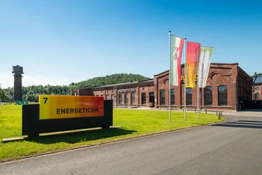 Haupteingang des Energeticon, ein ehemaliges Industriegelände mit Backsteingebäuden. Drei bunte Fahnen stehen einladend davor, und ein Turm ist links zu erkennen.