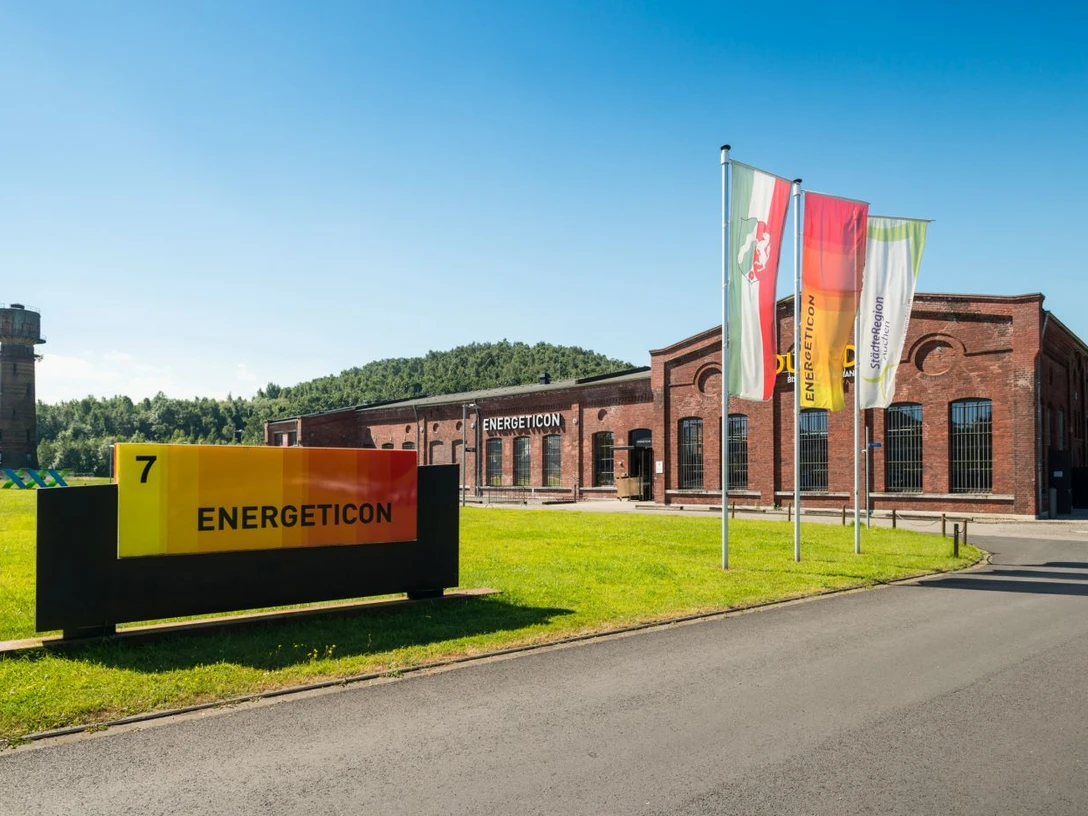 energeticon-haupteingang-dominik-ketz-2 Haupteingang des Energeticon, ein ehemaliges Industriegelände mit Backsteingebäuden. Drei bunte Fahnen stehen einladend davor, und ein Turm ist links zu erkennen.