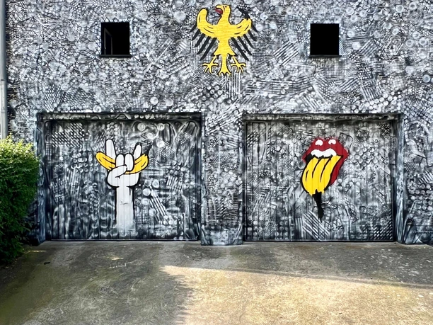 Spraygrammtechnik Bananensprayer Winckelmannstraße 11 1 Two garage doors are artistically painted on a gray, textured wall. Each door features a large graffiti: on the left, a white hand in pop art style, raising two fingers in a peace sign; on the right, a colorful illustration of the iconic "Stones" lip logo. Above it sits a yellow eagle, classic and royal, contrasting with the modern art below.