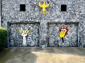 Spraygrammtechnik Bananensprayer Winckelmannstraße 11 Auf einer grauen, strukturierten Wand sind zwei Garagentore kunstvoll bemalt. Jedes Tor zeigt ein großes Graffiti: Links eine weiße Hand in Pop-Art-Stil, die zwei Finger in einem Friedenszeichen erhebt, rechts eine bunte Illustration des ikonischen "Stones"-Lippenlogos. Darüber thront ein gelber Adler, klassisch und royal, kontrastreich zur modernen Kunst darunter. Two garage doors are artistically painted on a gray, textured wall. Each door features a large graffiti: on the left, a white hand in pop art style, raising two fingers in a peace sign; on the right, a colorful illustration of the iconic "Stones" lip logo. Above it sits a yellow eagle, classic and royal, contrasting with the modern art below.