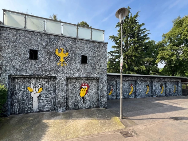 Spraygrammtechnik Bananensprayer Winckelmannstraße 11 2 Graffiti an einer grauen Fassade: Bananensymbole und Rolling-Stones-Mund illustrieren die Wand.